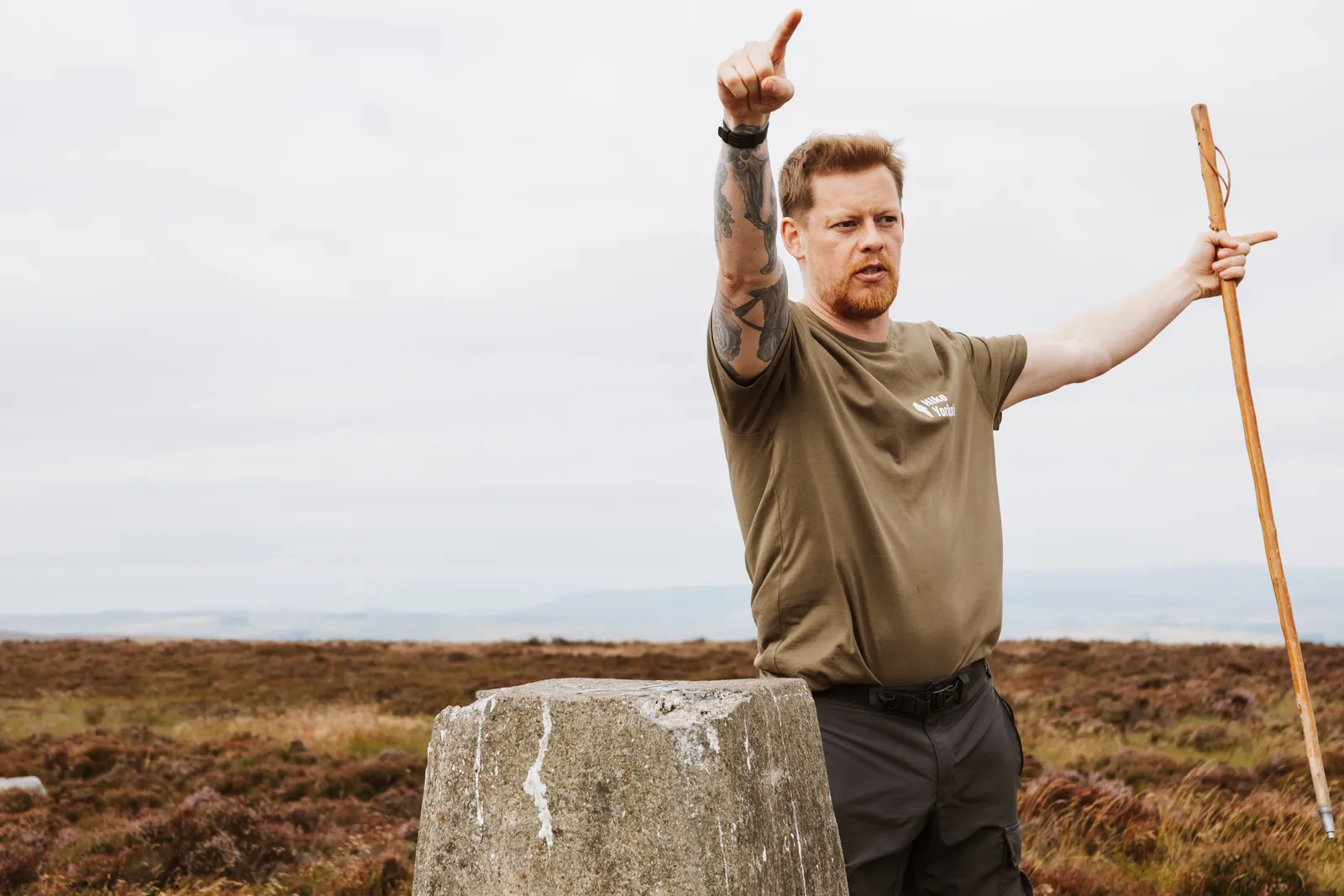 A Hike Yorkshire instructor pointing out a landmark beside a trig point on open moorland